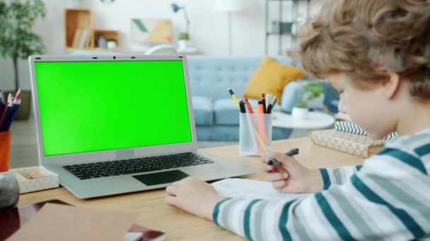 Smart kid learning online looking at green screen laptop and writing at home — Stock Video