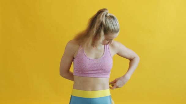 Linda chica en ropa deportiva midiendo la cintura con cinta métrica y sonriendo sobre fondo amarillo — Vídeos de Stock