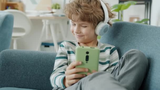 Cámara lenta de niño lindo con auriculares usando teléfono inteligente en casa — Vídeos de Stock
