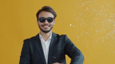 Sarı arka planda dans eden zarif takım elbiseli neşeli Hintli adam etrafta konfeti uçuşuyor.
