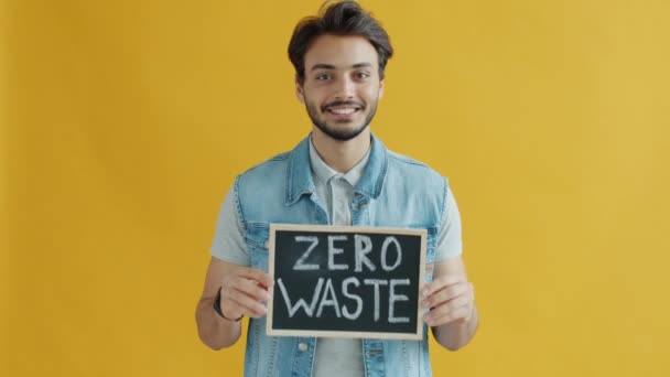 Movimento lento do cara de raça mista sorrindo segurando sinal Zero Waste no fundo de cor amarela — Vídeo de Stock