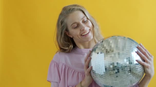 노란 배경 위에 서 있는 디스코 공을 들고 웃고 있는 명랑 한 젊은 여자의 모습 — 비디오