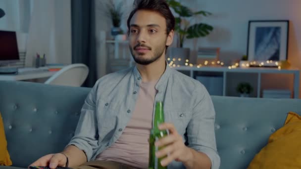 Radostný mladý muž sleduje televizi, směje se a pije pivo z láhve v temném domě v noci — Stock video