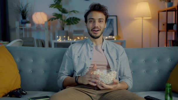 Портрет веселого ближневосточного человека, смеющегося, наслаждающегося фильмом по телевизору и поедающего попкорн дома ночью — стоковое видео