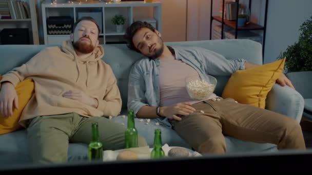 Két férfi barát tévét néz a kanapén fekve egy koszos lakásban éjszaka. — Stock videók