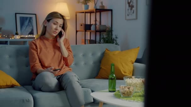 Ritratto di bella giovane donna che chiacchiera sul cellulare e guarda la TV a casa di notte — Video Stock