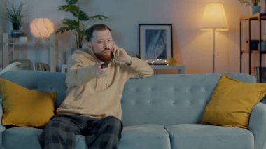 Rahat genç bir adam TV seyrediyor ve cep telefonuyla konuşuyor. Geceleri evdeki koltukta oturuyor. İletişim ve gençlik yaşam tarzı kavramı.