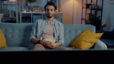 Korkmuş genç bir adam patlamış mısır yiyor ve gecenin karanlığında kanepede oturmuş gerilim filmi seyrediyor. Eğlence ve duygu konsepti.