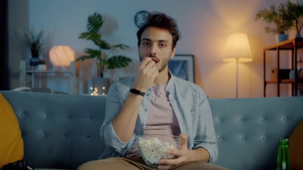 Портрет красивого парня, смотрящего телевизор, поедающего попкорн, напуганного по ночам дома — стоковое видео