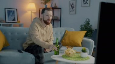 Yakışıklı bir öğrenci televizyon seyrediyor ve karanlık bir dairede şişeden bira içiyor.