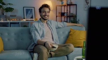 Neşeli erkek öğrenci gülerek ve patlamış mısır yiyerek karanlık odada televizyon izliyor.