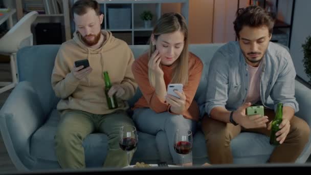 Διαφορετική ομάδα φίλων που χρησιμοποιούν smartphones πίνοντας αλκοόλ και βλέποντας τηλεόραση στο σπίτι το βράδυ — Αρχείο Βίντεο