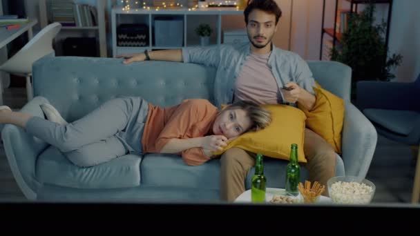 Ung kvinna tittar skrämmande film på TV med pojkvän liggande på soffan gömmer sig bakom kudde i mörkt rum — Stockvideo