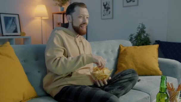 Портрет счастливого молодого человека, смотрящего телевизор, поедающего чипсы и пьющего пиво в квартире вечером. — стоковое видео