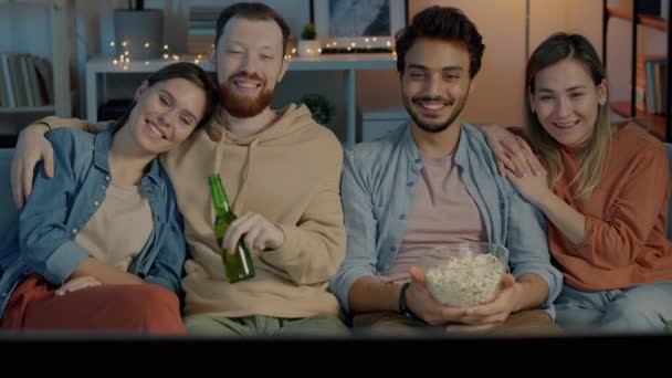 Счастливые друзья смотрят телевизор, наслаждаясь кукурузой и пивом в квартире по ночам — стоковое видео