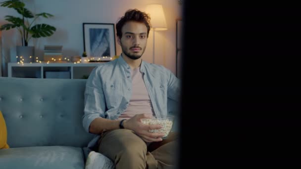 Эмоциональный ближневосточный мужчина смотрит триллер по телевизору и ест попкорн в освещенном доме — стоковое видео