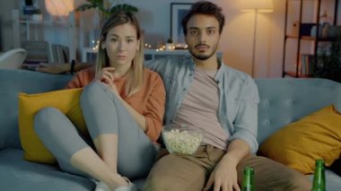 TV 'de korku filmi izleyen iki kişinin portresi. Koyu renkli bir dairede patlamış mısır yiyor.