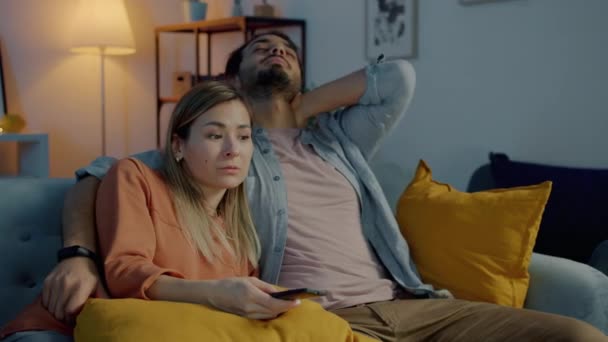 Сонная молодая пара смотрит телевизор, разговаривает и обнимается вечером в квартире — стоковое видео
