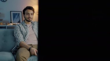 Orta Doğulu bir adam TV seyrediyor ve gülümsüyor. Elinde kumandayla karanlık bir dairede.