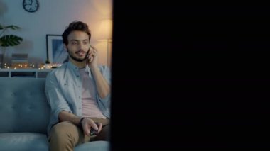 Çekici bir genç adam cep telefonuyla konuşuyor ve geceleri karanlık bir dairede televizyon izliyor.