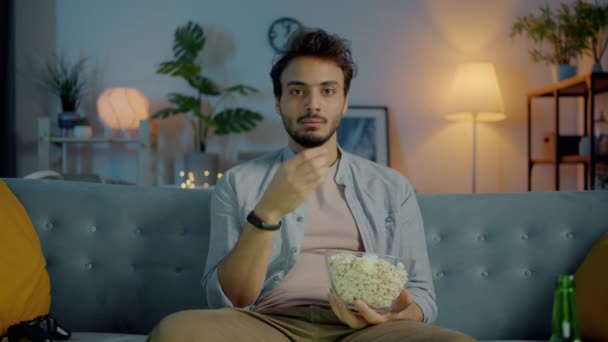 Портрет задумчивого ближневосточного человека, смотрящего телевизор, поедающего попкорн и думающего в темной квартире — стоковое видео