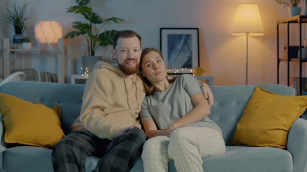 Älskade par i pyjamas tittar på TV hemma sitter på soffan och ler njuter av kvällen tillsammans — Stockvideo