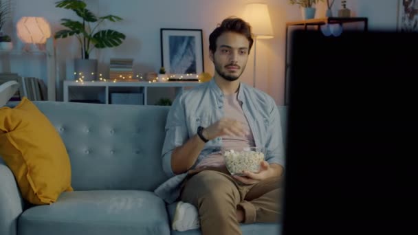 Feliz hombre de Oriente Medio comiendo palomitas de maíz y viendo la televisión relajándose en casa por la noche — Vídeo de stock