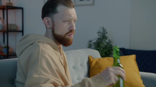 Emocjonalny młodzieniec ogląda straszny film w telewizji i pije piwo z butelki w oświetlonym domu — Wideo stockowe