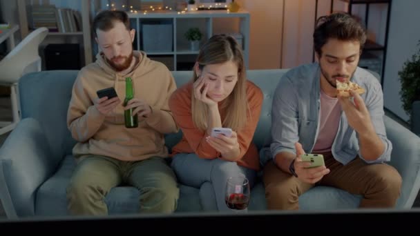 Amis multiculturels utilisant des smartphones boire de l'alcool manger de la pizza et regarder la télévision la nuit dans un appartement sombre — Video