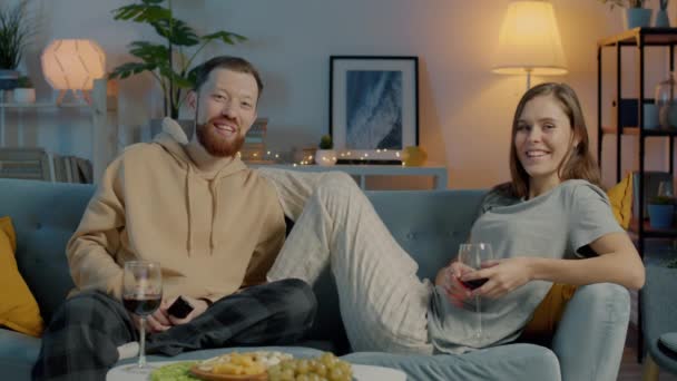 Mutlu genç bir çiftin portresi TV izliyor şarap içiyor kadeh tokuşturuyor ve karanlık evde gülümsüyor. — Stok video