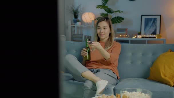 Wanita cantik birasial menonton TV tertawa dan minum dari botol di apartemen gelap — Stok Video