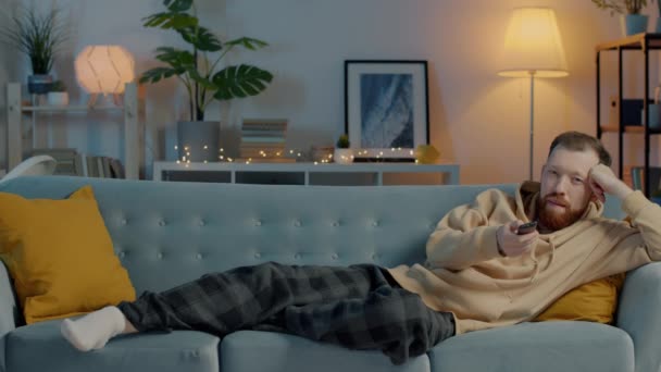 Zmęczony młodzieniec w domowych ciuchach oglądający telewizję wieczorem zasypiający na kanapie w domu — Wideo stockowe
