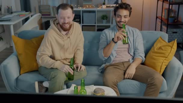 Ευτυχισμένοι νέοι άνδρες Καυκάσιος και Μέση Ανατολή βλέποντας τηλεόραση γελώντας μιλώντας και πίνοντας μπύρα στο σπίτι τη νύχτα — Αρχείο Βίντεο