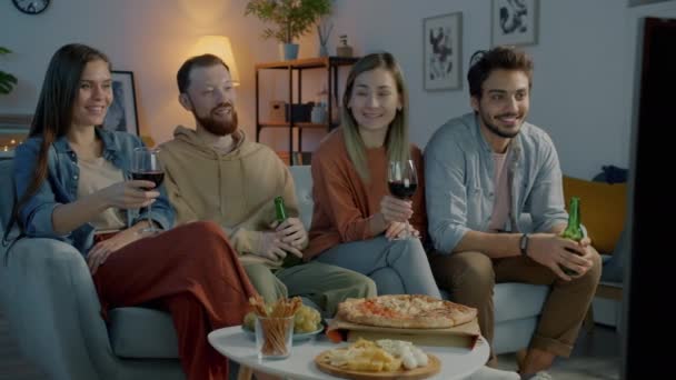 Grupa młodych ludzi oglądających telewizję pijących alkohol i jedzących pizzę w ciemnym mieszkaniu w nocy — Wideo stockowe
