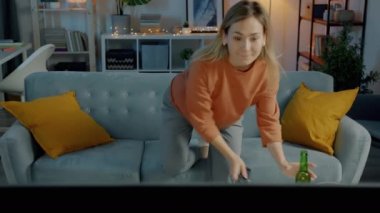 Ev kıyafetleri içinde çekici genç bir kadın televizyon izliyor ve karanlık bir dairede köpüklü içecek içiyor.