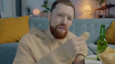 Çekici sakallı adamın portresi cips yiyor bira içiyor ve aydınlanmış evde televizyonda komik bir film izlerken gülüyor.