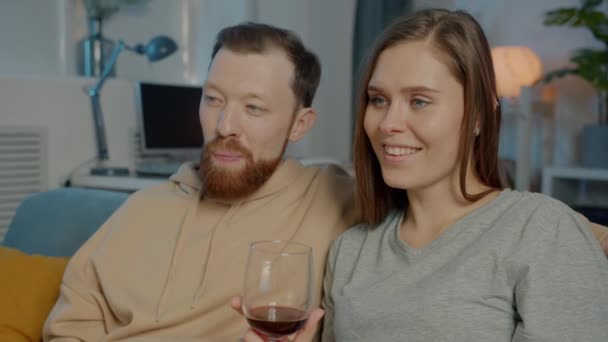 Молодая женщина смотрит телевизор с бойфрендом пьет красное вино и кормит мужчину сыром наслаждаясь вечером дома — стоковое видео