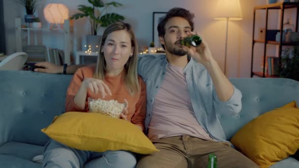 Glada par tittar sitcom skrattar äta popcorn och dricka öl i lägenheten på natten — Stockvideo