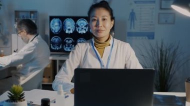 Beyaz cüppeli kadın pratisyen doktor portresi karanlık hastane ofisinde dizüstü bilgisayarla oturuyor.