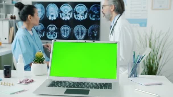 Chroma key green screen laptop with doctor and nurse in uniform discussing MRI scans in background — Stock Video