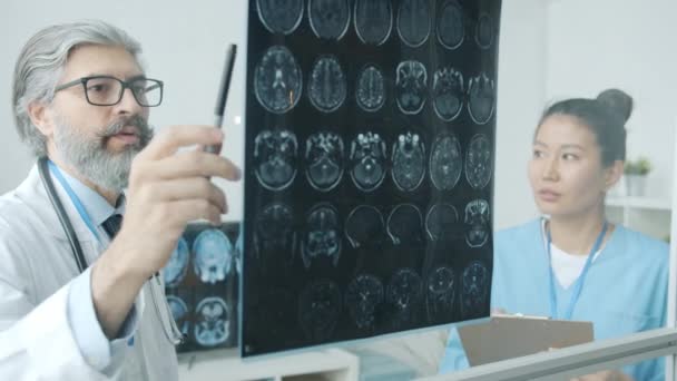 MRI脳画像を解析する白衣の医師、診療所で働く頭蓋骨の看護師と話す医師 — ストック動画