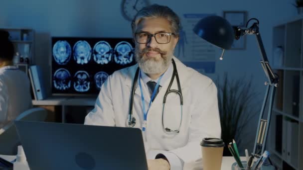 Portret dojrzałego mężczyzny w białym płaszczu siedzącego przy biurku w ciemnym gabinecie szpitalnym — Wideo stockowe