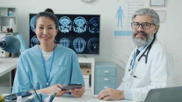 Arzt und Krankenschwester sitzen lächelnd am Arbeitsplatz im Krankenhaus und blicken in die Kamera — Stockvideo
