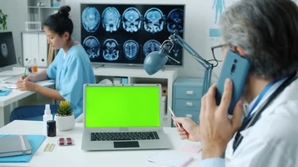 Médico falando no telefone celular e olhando para tela verde chroma laptop chave enquanto enfermeira escrevendo em segundo plano. — Vídeo de Stock