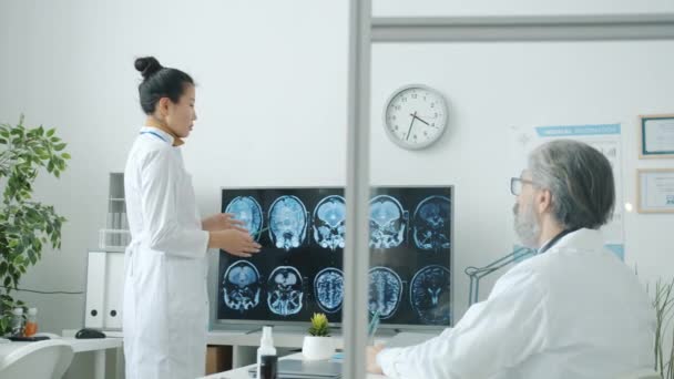 Neurólogo Asiática dama en blanco abrigo discutir MRI imágenes con macho colega caucásico hombre — Vídeos de Stock