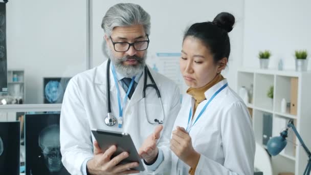 Médicos masculinos e femininos olhando para a tela do tablet conversando e sorrindo dentro de casa na clínica moderna — Vídeo de Stock