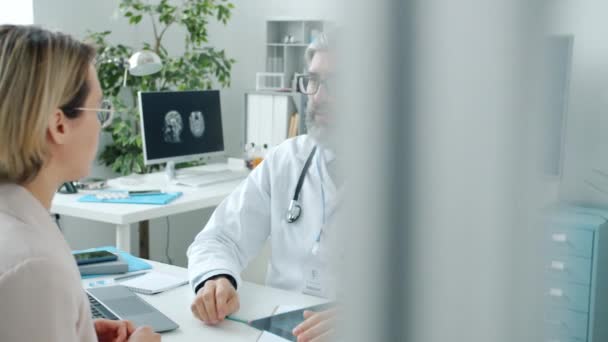 Male doctor discussing disease with young woman showing MRI scan and talking in clinic — Stock Video