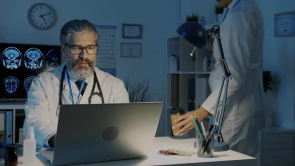 Doctor cansado recibiendo taza de café de una colega bebiendo trabajando en el hospital tarde en la noche — Vídeo de stock