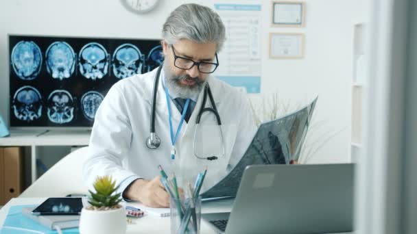 Doświadczony lekarz badający obraz rezonansu magnetycznego i korzystający z maszynopisania komputerowego pracujący w szpitalu — Wideo stockowe