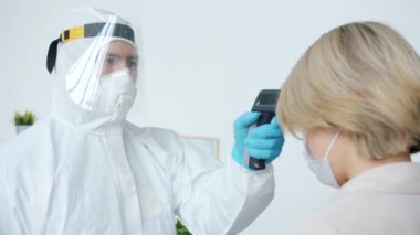 Koruyucu kostümlü doktor vücut ısısını kontrol ediyor. Kadın hastayı dokunmadan termometreyle muayene ediyor.